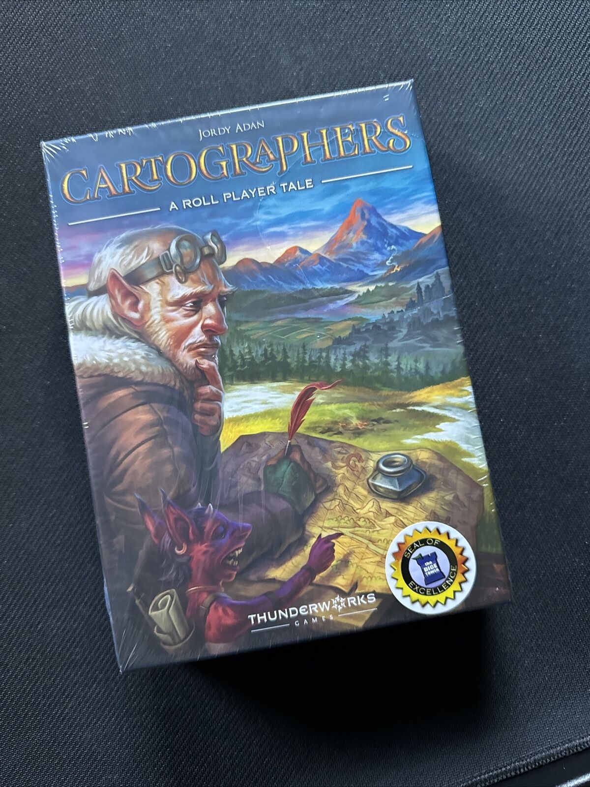 A photo of the board game "Cartographers: A Roll Player Tale" by Jordy Adan, sealed in plastic wrapping. The cover features an illustration of a bearded man with a pair of goggles on his head looking to the side and a red, fox-like creature holding a quill, both over a background depicting a fantasy landscape with mountains. The game is produced by Thunderworks Games and has a "Seal of Excellence" sticker from The Dice Tower.