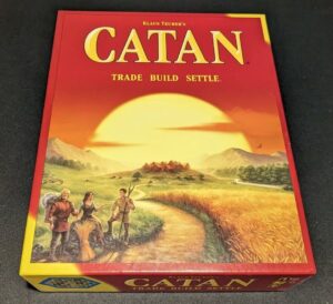 A photo of the board game "Klaus Teuber's CATAN" box with the tagline "Trade Build Settle" featuring artwork of three people on a landscape at sunset.