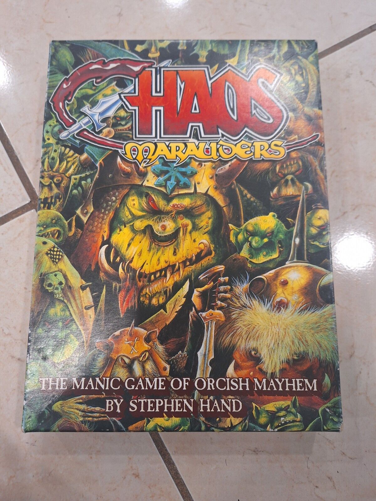 A board game titled "Chaos Marauders" with the tagline "THE MANIC GAME OF ORCISH MAYHEM BY STEPHEN HAND" is lying on a tiled floor. The cover features colorful artwork of cartoonish orc characters and fantasy elements.