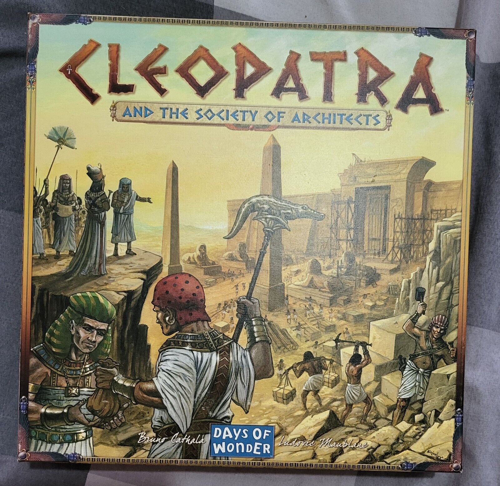 Board game cover for "Cleopatra and the Society of Architects" featuring illustrated ancient Egyptian characters and construction scene with pyramids and pillars in the background.