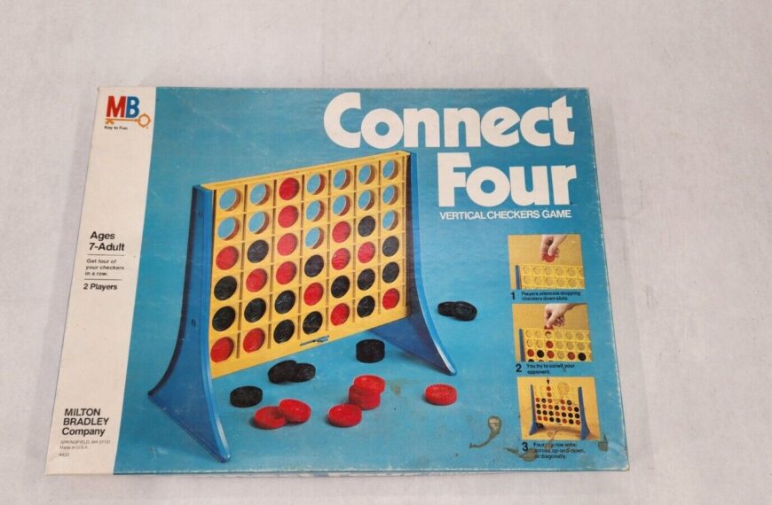 The image shows a Connect Four board game box with a visible playing grid standing upright, scattered red and black game pieces in front, and instructions on the side of the box.