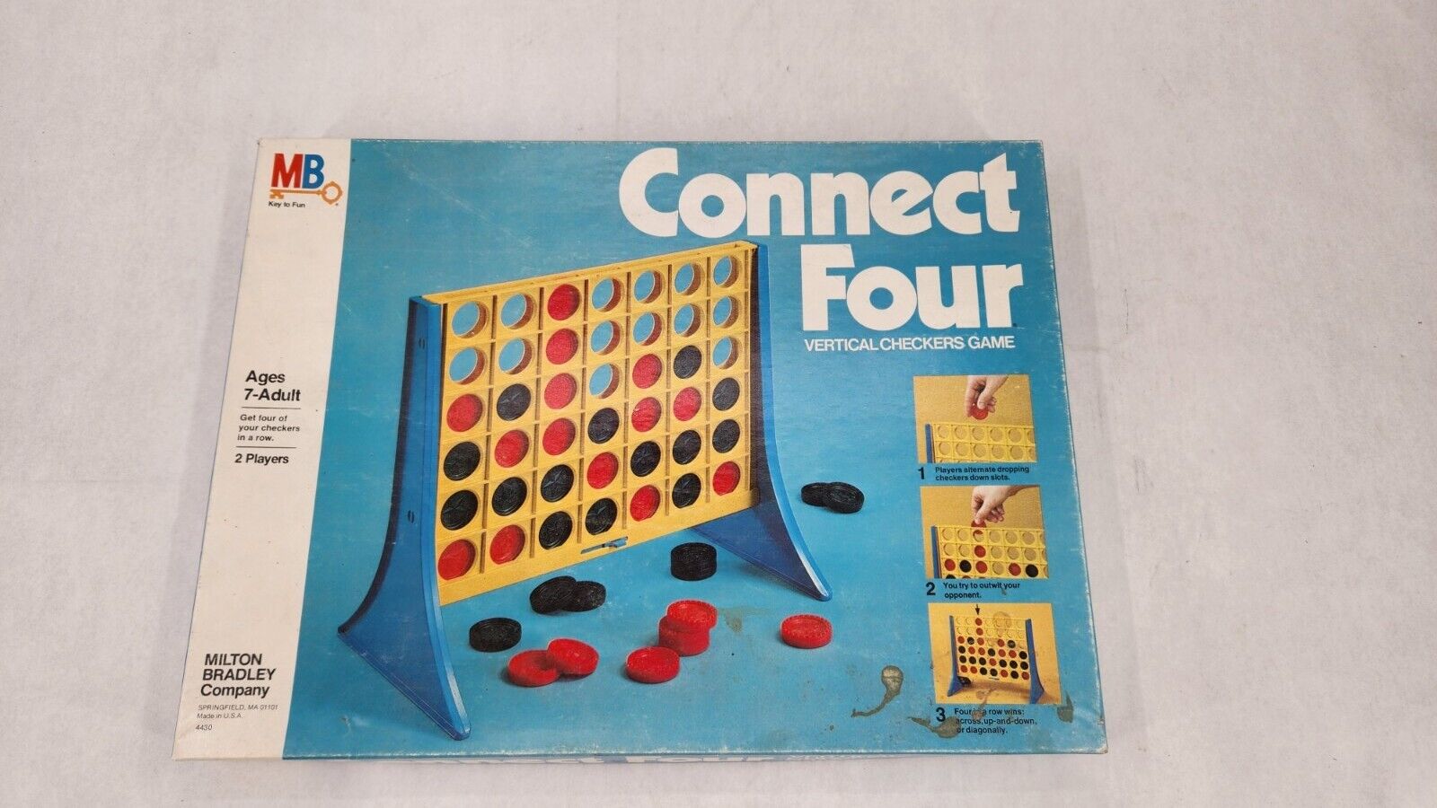 The image shows a Connect Four board game box with a visible playing grid standing upright, scattered red and black game pieces in front, and instructions on the side of the box.