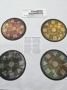 Four round game coasters with different color schemes labeled "Cosmic Coasters" alongside instructions on how to use them for a game about teleportation combat.