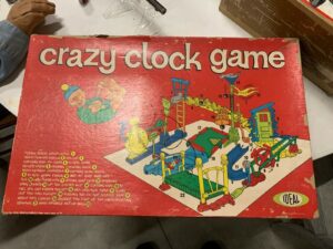 Vintage "Crazy Clock Game" box with illustrated game setup and a sequence of events, showing a clown and various mechanisms, on a red background. The IDEAL logo is in the bottom right corner. A hand is visible in the top left corner.
