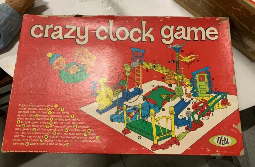 Vintage "Crazy Clock Game" box with illustrated game setup and a sequence of events, showing a clown and various mechanisms, on a red background. The IDEAL logo is in the bottom right corner. A hand is visible in the top left corner.