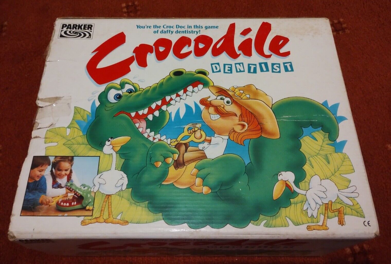 A damaged game box for "Crocodile Dentist" by Parker, featuring an illustration of a cartoon crocodile being treated by a dentist, with a whimsical jungle background and a small photo of two children playing the game in the corner.