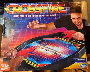 Box cover of the Crossfire board game, featuring intense graphics with two players holding red plastic guns aiming at game pieces on the board, with the slogan "Blast fast to win in this rapid-fire game!" Visible age recommendation is 7+ and a warning label for choking hazard. The Hasbro Gaming logo is in the bottom right corner.