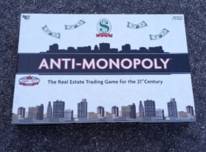 A board game called "ANTI-MONOPOLY" with the tagline "The Real Estate Trading Game for the 21st Century" on its cover, which also displays a city skyline, the logo "SINCE 1977 CLASSIC," money illustrations, and text indicating it is for ages 8 & up and 2 to 6 players.