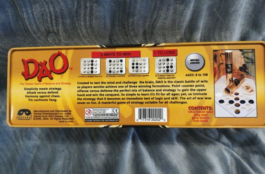 Image of a DAO strategy board game box describing the game as a "Classic Game of Balance and Strategy," suitable for ages 8 to 108 with a Mensa Award badge. The box illustrates three ways to win and one way to lose, contents of the game including a travel tube, play mat, gem stones, and rule book, and a background image of the game components set up for play.