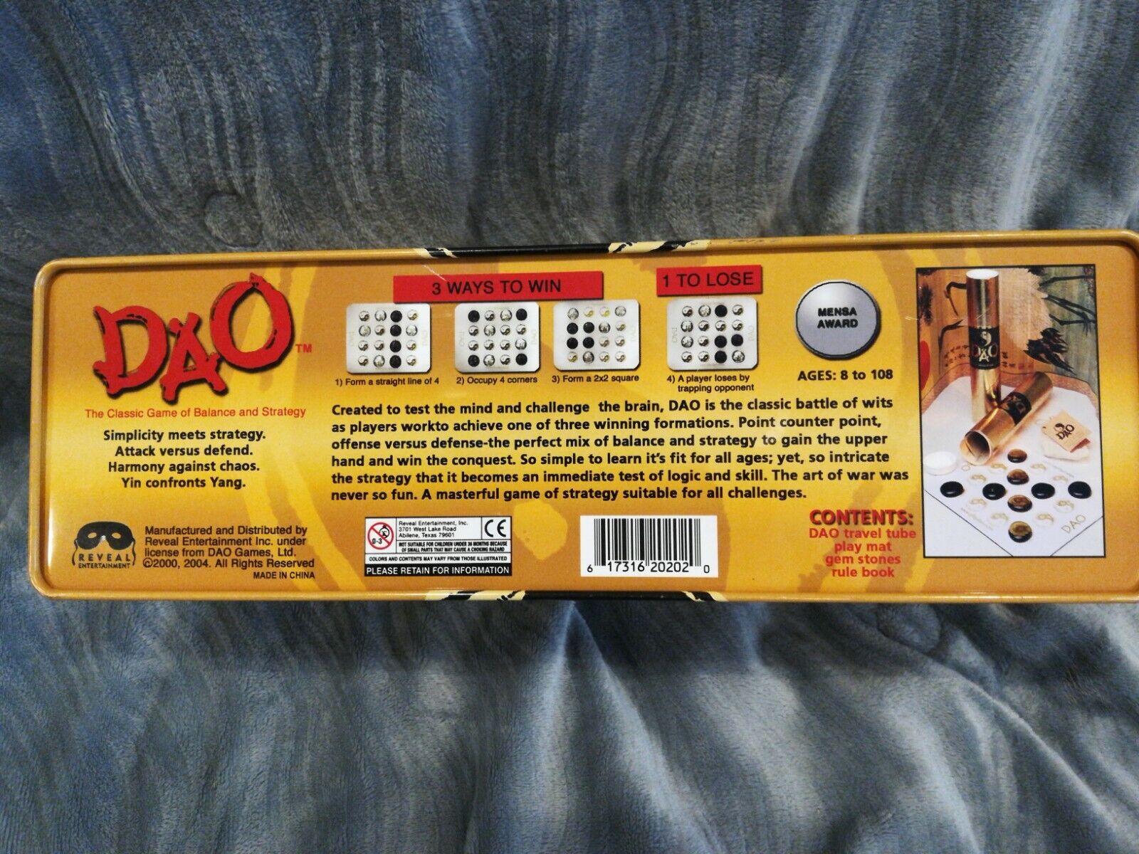 Image of a DAO strategy board game box describing the game as a "Classic Game of Balance and Strategy," suitable for ages 8 to 108 with a Mensa Award badge. The box illustrates three ways to win and one way to lose, contents of the game including a travel tube, play mat, gem stones, and rule book, and a background image of the game components set up for play.