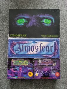 Alt text: A collection of "Atmosfear" board game boxes, including "The Harbingers" with glowing green eyes on the cover, and other editions featuring haunting imagery and promotional labels. The boxes appear slightly worn.