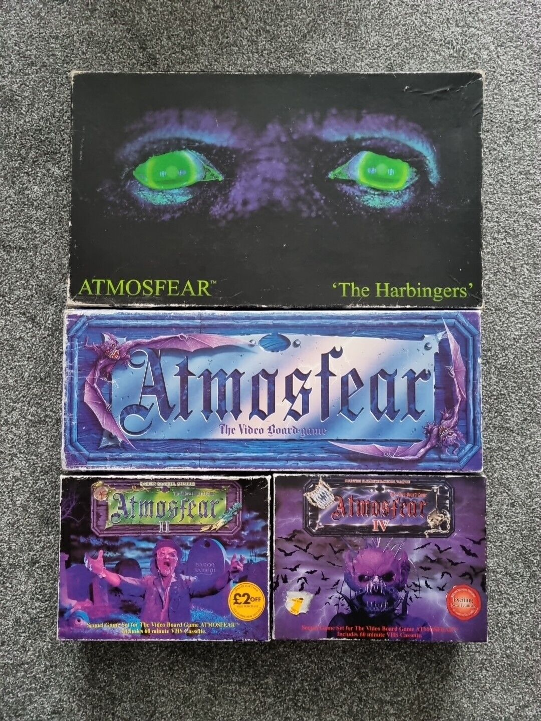 Alt text: A collection of "Atmosfear" board game boxes, including "The Harbingers" with glowing green eyes on the cover, and other editions featuring haunting imagery and promotional labels. The boxes appear slightly worn.