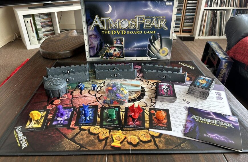 The image shows "Atmosfear: The DVD Board Game" set up and ready to play on a wooden table with the game's box visible in the background. The board features various character pieces, a well of Fears, numbered keys, dice, and a stack of cards. The interior is visible with shelves of DVDs and a couch in the background.