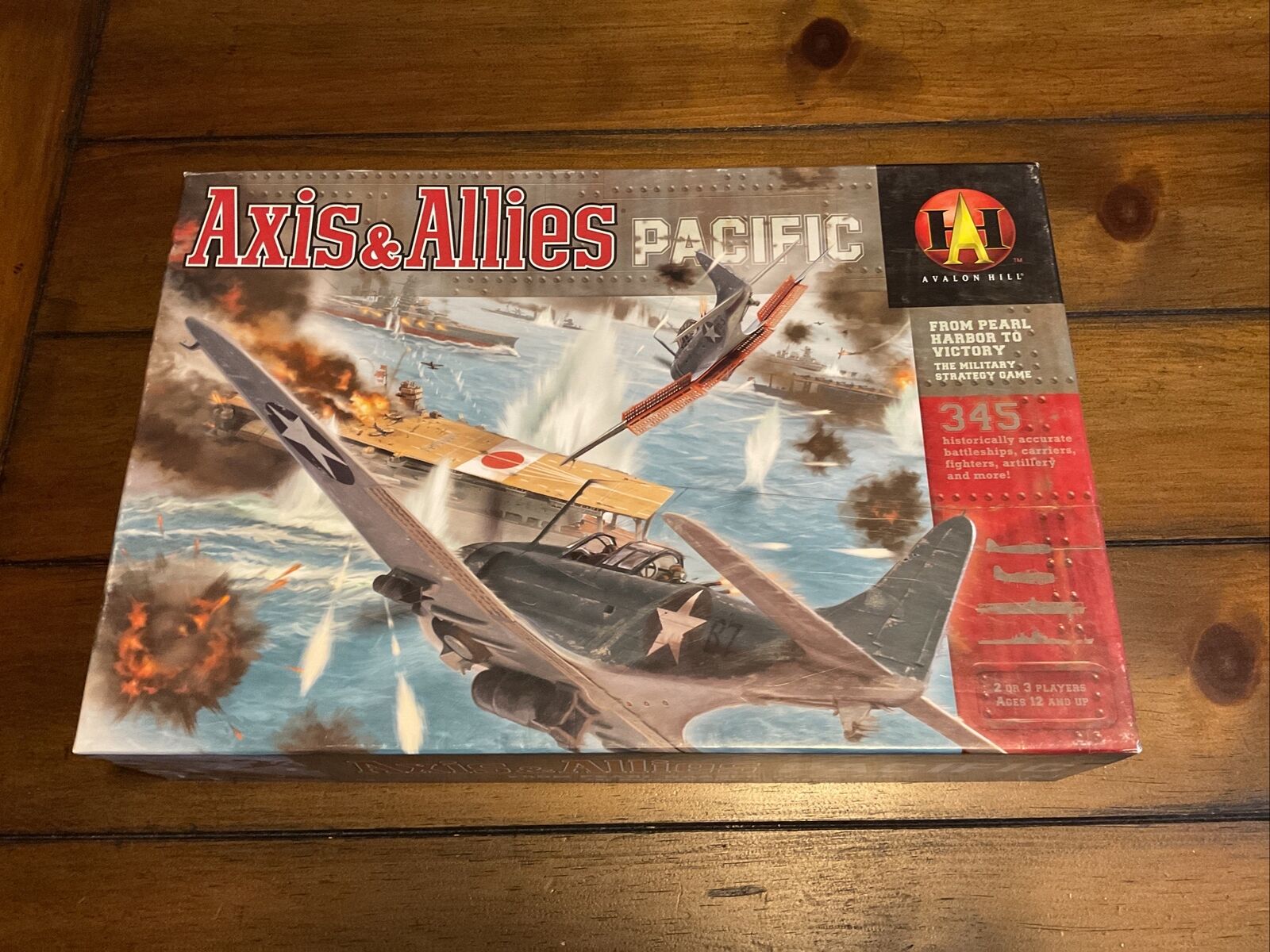 A board game titled "Axis & Allies Pacific" by Avalon Hill, depicting World War II imagery with planes, ships, and explosions, lying on a wooden surface.