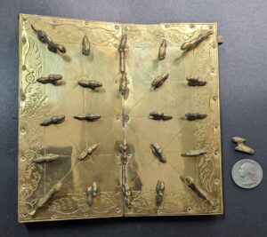 An ornate, brass-colored binding with engraved floral patterns and clasps shaped like turtles, displayed on a dark surface next to a coin for scale.