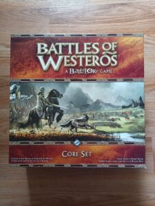 A board game named "Battles of Westeros: a Battlelore Game, Core Set" is placed on a wooden surface. The artwork on the box features medieval-style battle scenes with knights and soldiers in a fantasy landscape.