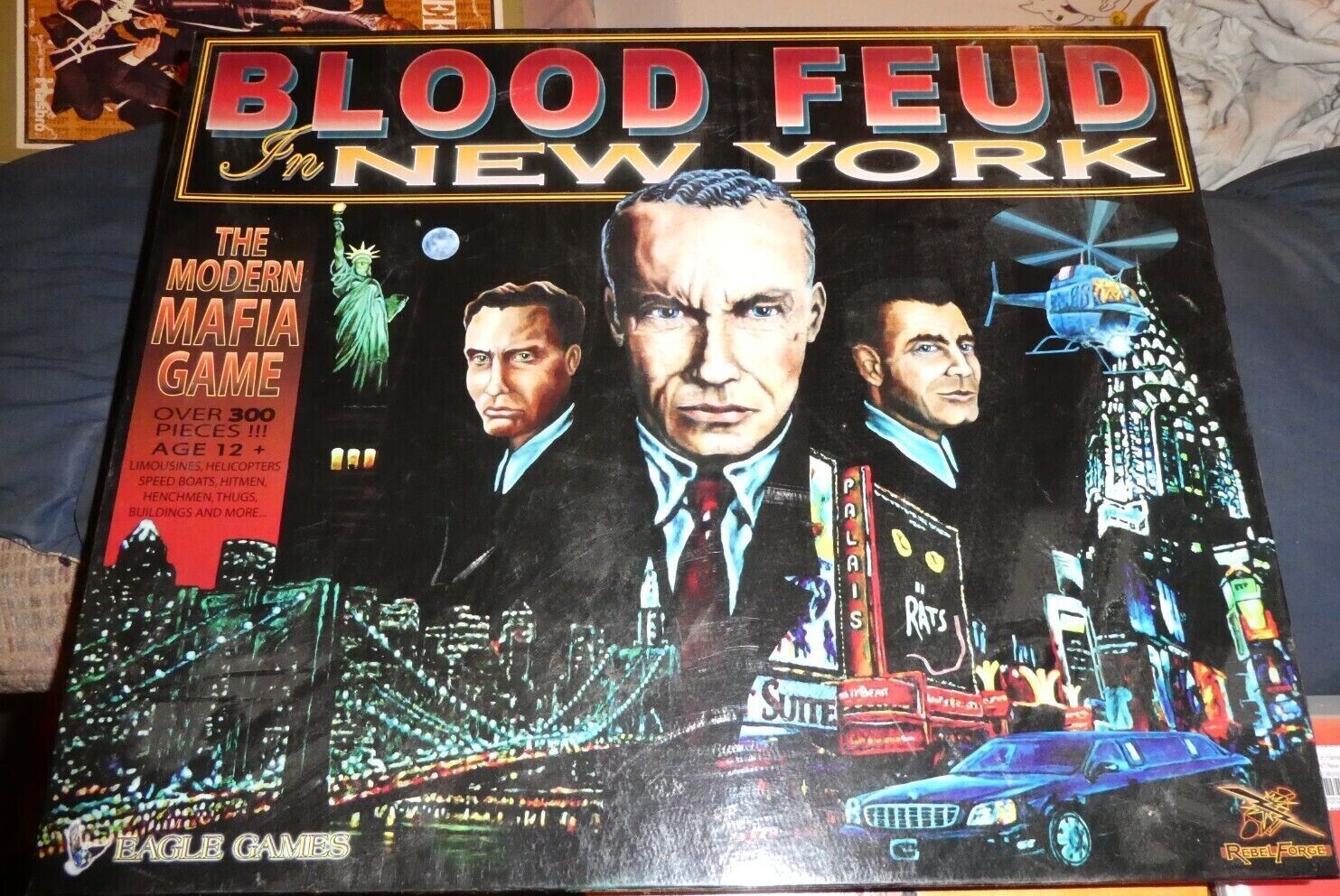 Board game cover art for "Blood Feud in New York" featuring a nighttime scene with illustrated mobster characters, a helicopter, limousine, and New York City skyline with the Statue of Liberty. Text highlights the game's aim and includes phrases such as "The Modern Mafia Game" and "Over 300 Pieces". The game is for ages 12 and up and is produced by Eagle Games.