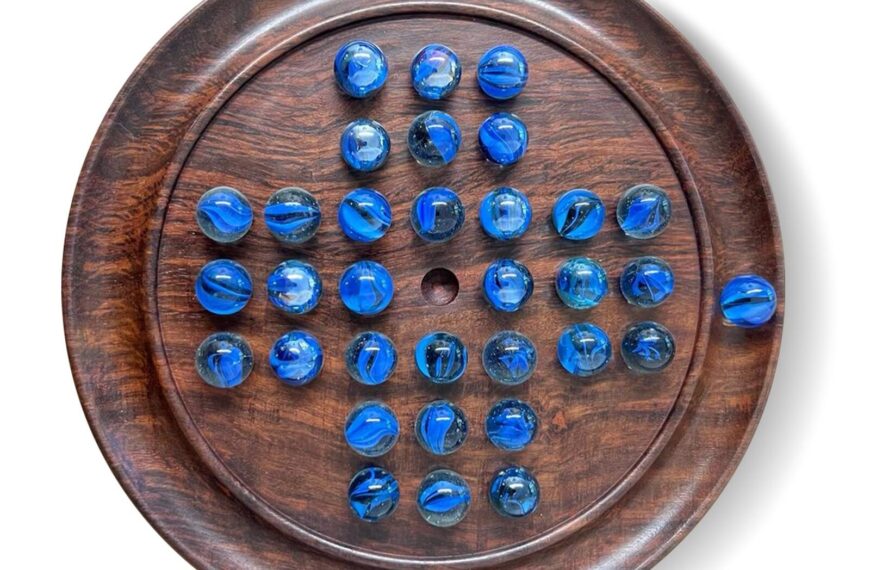 A wooden solitaire board with blue marbles arranged in the starting pattern, with the center space empty, isolated on a white background.
