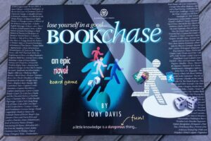 An advertising poster for "Bookchase," an epic novel board game by Tony Davis, with text and images including game pieces, dice, and a list of classic books scattered across the background. The tagline reads, "Lose yourself in a good... Bookchase" and "a little knowledge is a dangerous thing... fun!"