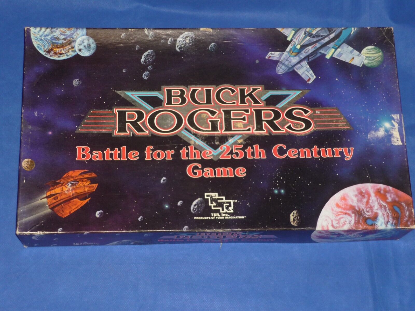 A Buck Rogers Battle for the 25th Century board game box on a blue fabric surface, featuring space-themed artwork with planets, asteroids and spacecraft.