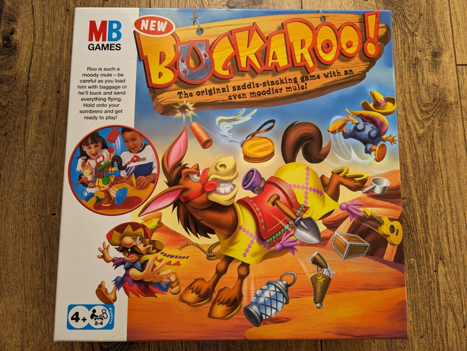 Board game "Buckaroo!" packaging with an animated bucking mule, flying objects, and a Western theme. The game's slogan "The original saddle-stacking game with an even moodier mule" is displayed, along with the MB Games logo and an image of two children playing the game. The packaging notes a 4+ age rating and is for 2-4 players.