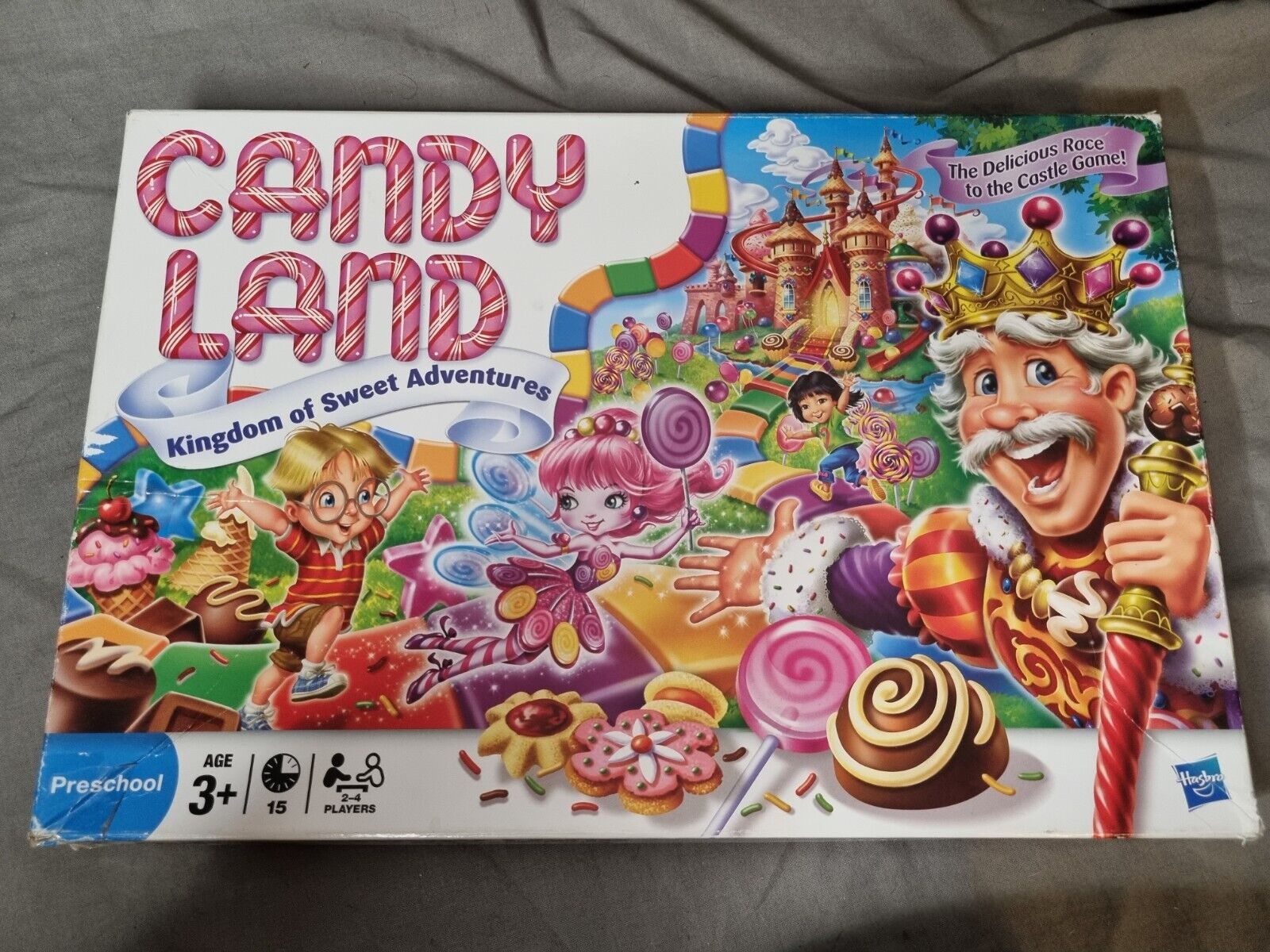 A "Candy Land" board game box featuring cartoon characters, a colorful candy-themed path, a lollipop forest, and a castle in the background. The text on the box reads "Kingdom of Sweet Adventures" and "The Delicious Race to the Castle Game!" Age recommendation is 3+, for 2-4 players, by Hasbro.