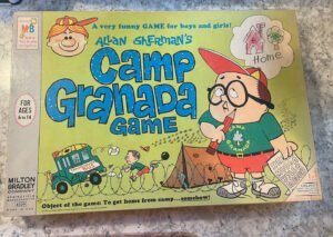 Vintage "Allan Sherman's Camp Granada Game" board game box cover featuring cartoon illustrations including a boy in camp attire with a thought bubble of a house labeled "Home," another child entangled in telephone wire, and a bus with "Camp Bunks" written on the side. Text on the box reads "A very funny GAME for boys and girls!," "For ages 6 to 14," "Milton Bradley Company Springfield Massachusetts," and "Object of the game: To get home from camp... somehow." The box shows signs of wear and a handwritten note.