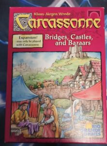 Box cover of "Carcassonne: Bridges, Castles, and Bazaars" expansion with medieval-themed illustration, including characters trading goods in a market and a castle in the background.