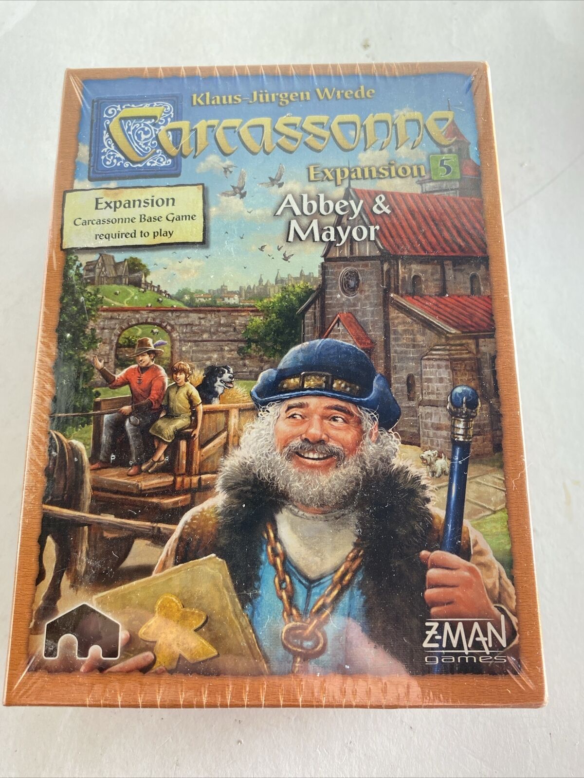 The image shows the box cover of the "Carcassonne Expansion 5: Abbey & Mayor" board game by Klaus-Jürgen Wrede. The artwork features a medieval setting with a smiling man in the foreground holding a blue staff and two game tile pieces, and a scene with a wagon and its driver in the background beside a stonework building. The Z-Man Games logo is present on the bottom right.