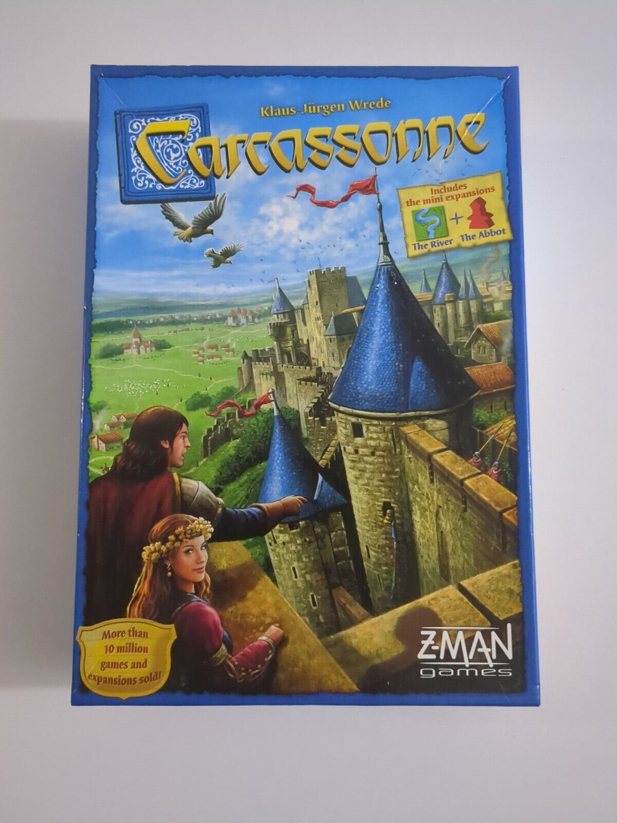 A photo of the board game "Carcassonne" by Klaus-Jürgen Wrede. The cover features a medieval landscape with a castle and two illustrated figures, a man and a woman, looking over a rampart. A crest with the game's name flies above them like a banner, with birds in the sky. Text on the cover states "Includes the mini expansions The River The Abbot" and "More than 10 million games and expansions sold." The Z-Man Games logo is in the bottom right corner.