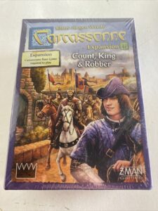 A sealed copy of the board game "Carcassonne Expansion 6: Count, King & Robber" on a tabletop, with artwork depicting medieval characters including a mounted knight and a person in a purple cloak. The text on the cover indicates it is an expansion and requires the Carcassonne base game to play. The game is by Klaus-Jürgen Wrede and published by Z-Man Games.