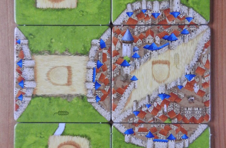 Six square tiles from a board game arranged in a 2x3 grid on a wooden surface, depicting segments of a medieval town with houses, a river, and green fields, with some tiles featuring symbols. Blue meeples are placed on certain tiles, indicating player moves or actions.