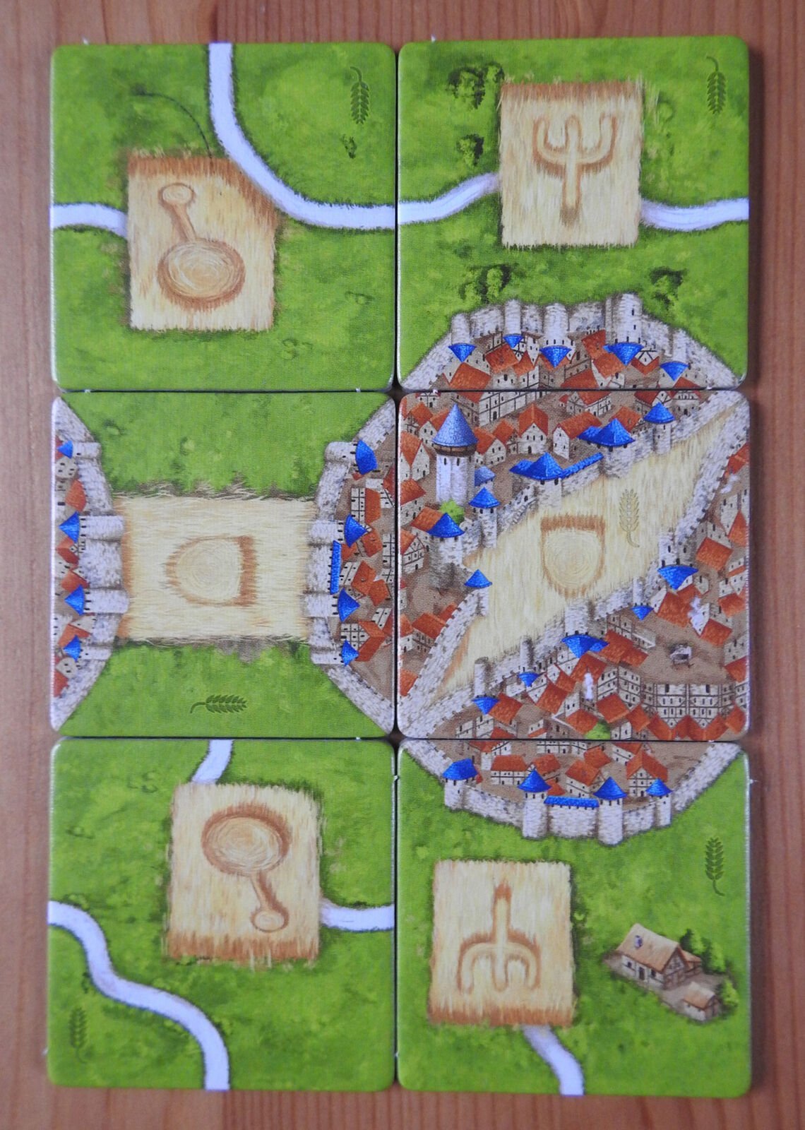 Six square tiles from a board game arranged in a 2x3 grid on a wooden surface, depicting segments of a medieval town with houses, a river, and green fields, with some tiles featuring symbols. Blue meeples are placed on certain tiles, indicating player moves or actions.