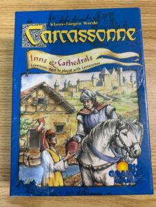 The image shows the cover of a board game named "Carcassonne: Inns & Cathedrals" by Klaus-Jürgen Wrede, published by Rio Grande Games. It features an illustration of a knight on horseback receiving something from a villager against a background of medieval buildings, with "Expansion: must be played with Carcassonne" written under the title.