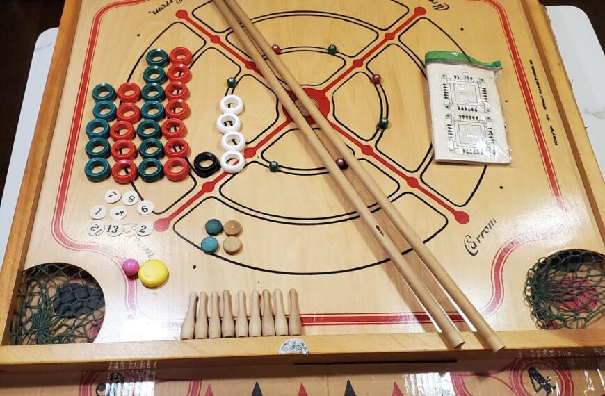 A Carrom board with coins, striker, and cues arranged for play, with a scorecard and an unused net pocket visible on the side.