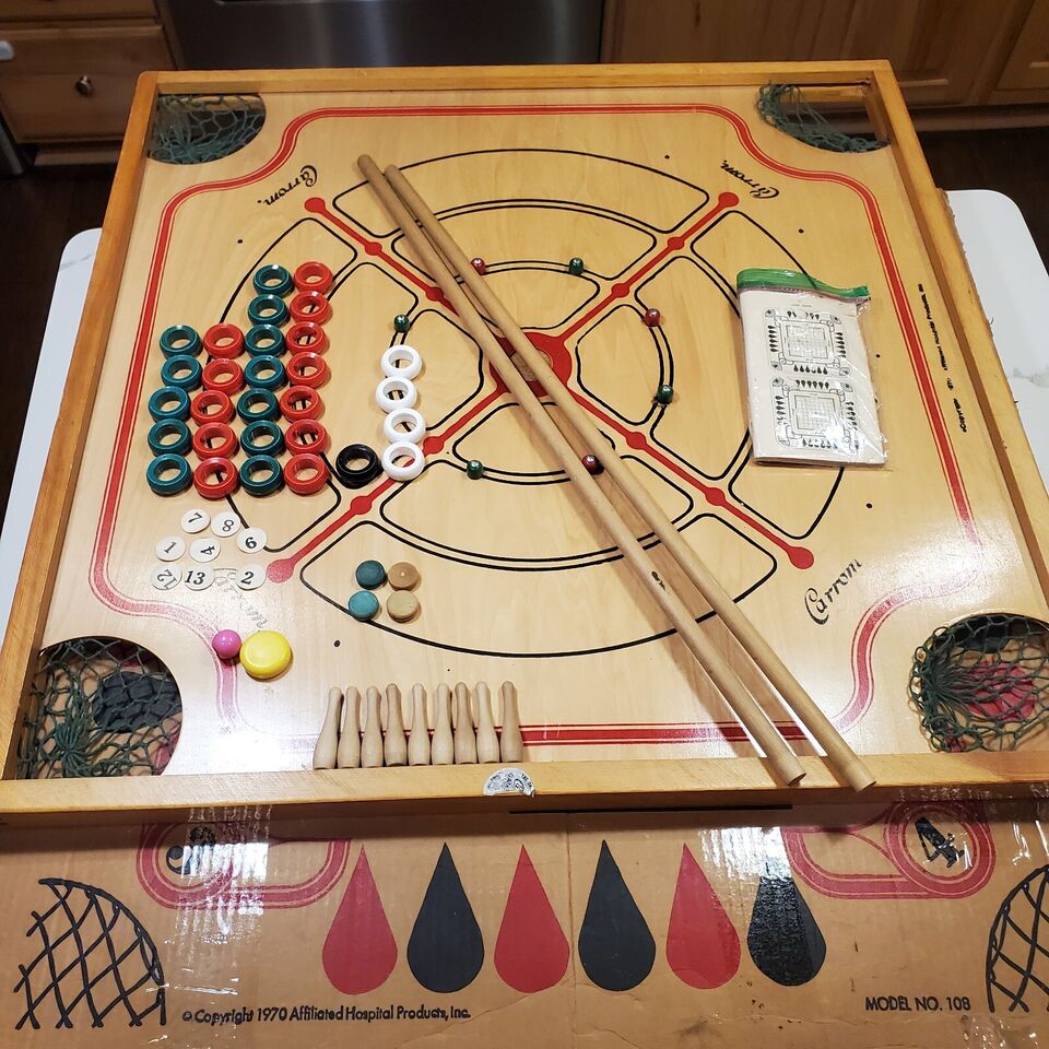 A Carrom board with coins, striker, and cues arranged for play, with a scorecard and an unused net pocket visible on the side.