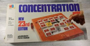 Box of the "Concentration" board game, labeled as "New 23rd Edition" by Milton Bradley, showing an image of the game with numbered tiles, for ages 10 to adult and designed for 2-5 players, placed on a wooden surface.