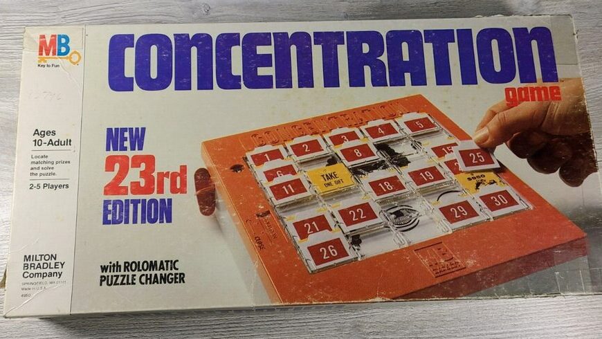 Box of the "Concentration" board game, labeled as "New 23rd Edition" by Milton Bradley, showing an image of the game with numbered tiles, for ages 10 to adult and designed for 2-5 players, placed on a wooden surface.