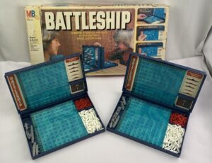 A vintage Battleship board game with its box in the background, and two foldable game boards open in the foreground showing blue grids and compartments with white and red pegs and miniature ship pieces.