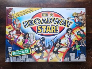Board game cover for "Be A Broadway Star!" featuring colorful cartoon illustrations of various characters partaking in Broadway-related activities with a large title in the center and a Broadway street sign at the bottom. The cover also includes the text "Can you make it to the Broadway Hall of Fame?" and information about the suitable age range and number of players.