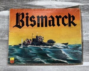 A worn vintage board game box titled "Bismarck" featuring an illustration of a battleship at sea with explosions around it, set against a yellow-to-black gradient background. The Avalon Hill Company logo is at the bottom left.