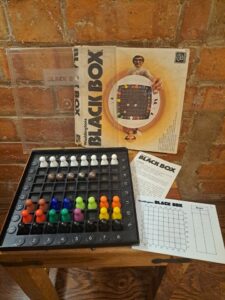 A vintage "Black Box" board game is arranged for play on a wooden stool against a brick wall. The game components include a black game board with colorful pegs, clue cards, and metallic balls. The game's box art features a man holding the board, and an instruction sheet is visible next to the box.