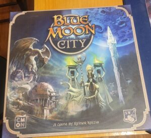 A board game box titled "Blue Moon City," featuring a fantastical illustration with a winged creature, a robed figure holding a candelabra, and an ancient cityscape, designed by Reiner Knizia. The publisher logos for CMON and SWG are visible in the corners.