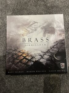 A photo of the board game "Brass: Birmingham" cover with artwork depicting an industrial landscape, featuring the names Matt Tolman, Martin Wallace, and Gavan Brown, and the Roxley games logo.