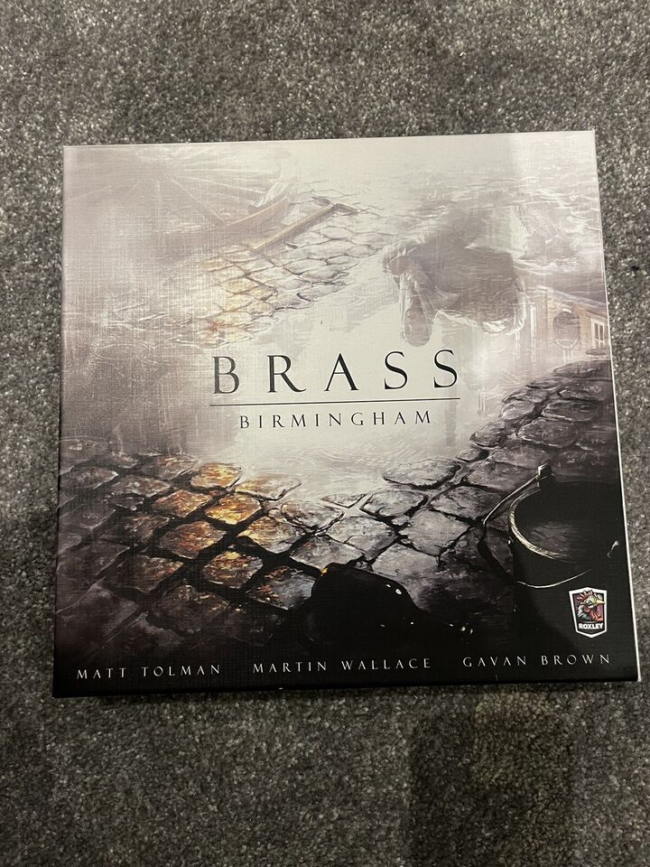 A photo of the board game "Brass: Birmingham" cover with artwork depicting an industrial landscape, featuring the names Matt Tolman, Martin Wallace, and Gavan Brown, and the Roxley games logo.