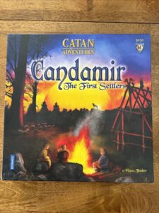 Board game box for "Catan Adventures: Candamir – The First Settlers" depicting a twilight scene with silhouetted trees, settlers around a campfire, and a partially constructed wooden structure against an orange sky. The Kosmos logo is in the bottom left corner and the designer's name, Klaus Teuber, is at the bottom right.