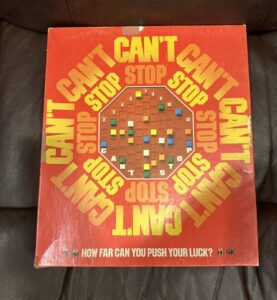 A worn board game box titled "CAN'T STOP" with a red and orange color scheme lying on a leather surface, and the tagline "HOW FAR CAN YOU PUSH YOUR LUCK?" at the bottom.