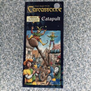 Box cover of "Carcassonne: Catapult", an expansion for the Carcassonne board game, featuring illustrated medieval characters operating a catapult and others juggling on stilts, against a backdrop of wooden structures and a crowd of onlookers.
