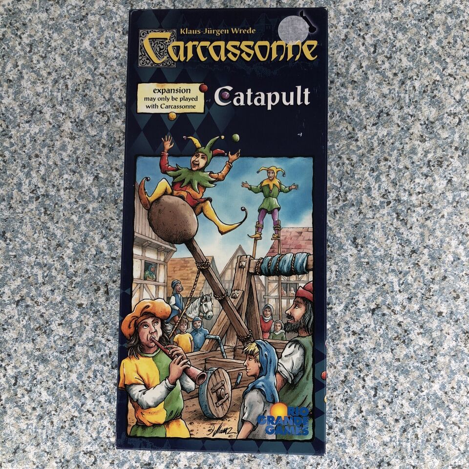Box cover of "Carcassonne: Catapult", an expansion for the Carcassonne board game, featuring illustrated medieval characters operating a catapult and others juggling on stilts, against a backdrop of wooden structures and a crowd of onlookers.