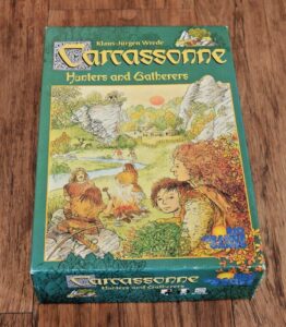 A board game box for "Carcassonne: Hunters and Gatherers" on a wooden surface, featuring illustrated prehistoric people and a lush landscape.
